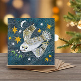 A square greeting card featuring an illustrated snowy owl flying across a dark blue starry night sky, with a crescent moon in the background  and mistletoe in the owl's beak.