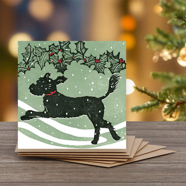 A square Christmas greeting card featuring a black dog jumping under a holly garland.