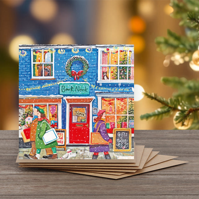 A square Christmas greeting card featuring a colorful painting of a cozy storefront scene with people, a wreath, and holiday decorations.