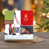 A square Christmas greeting card with a painting  of a small brown dog wearing a colorful striped Christmas sweater.