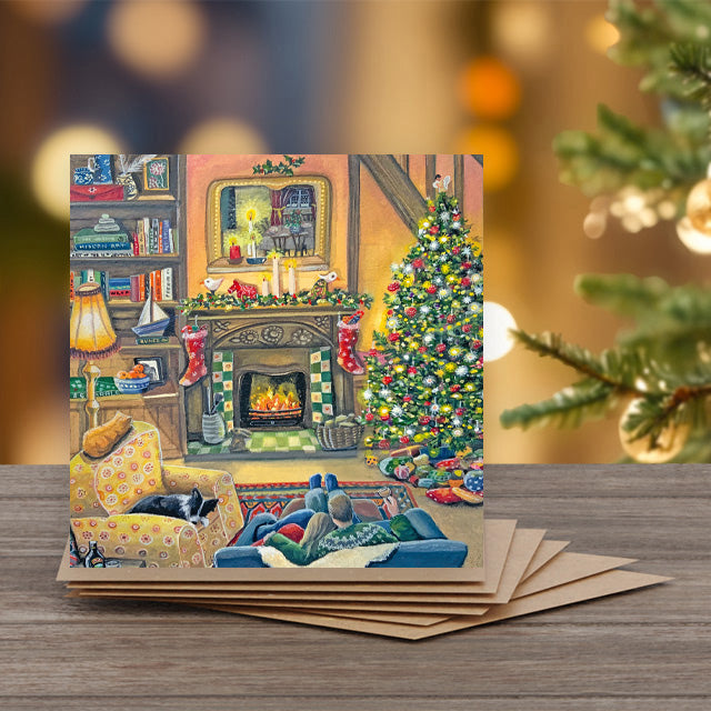 A square greeting card featuring a cozy Christmas living room scene with a fireplace, decorated tree, bookshelves, and people lounging on a couch.